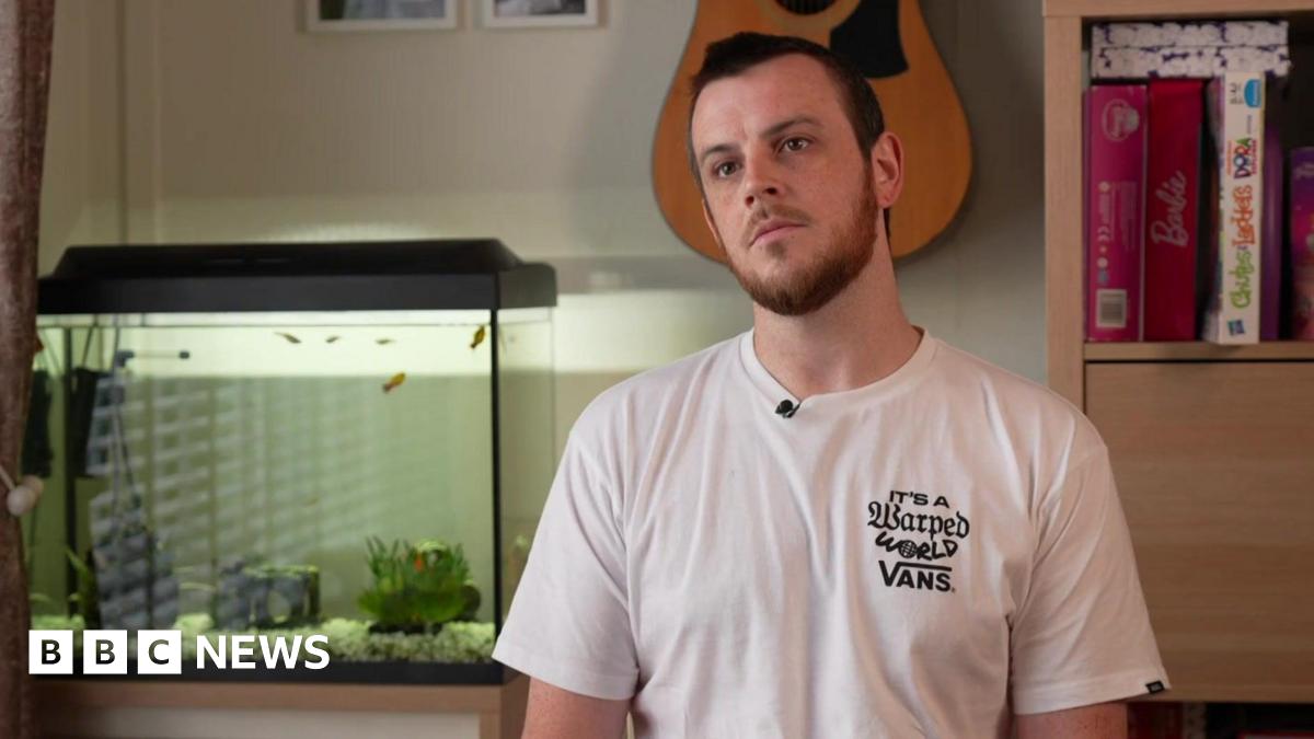 Marcus Johnson wears a white tee-shirt sitting in a room with a fish tank and a guitar on the wall.