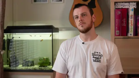 BBC Marcus Johnson wears a white tee-shirt sitting in a room with a fish tank and a guitar on the wall.