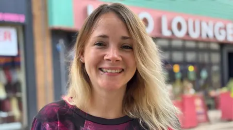Woman with shoulder-length blonde hair and wearing a purple and black long sleeved top smiling on Wolverhampton's high street