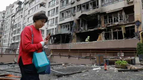 EPA A woman wearing a red jumper and holding her phone walks past a bombed-out residential building in Kyiv on 21 July.
