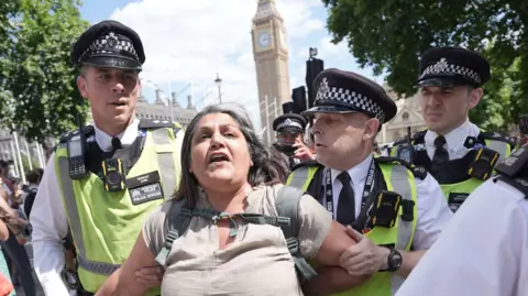 PA Media A woman is led away by police officers as supporters of Palestine Action take part in a protest in Parliament Square, Westminster