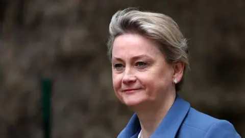 EPA A close up of Yvette Cooper as she walks past some bushes. She is smiling slightly and wearing a blue jacket and white leaf-shaped earrings. Her hair is cropped short.