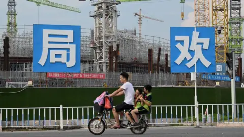 Getty Images People commute in front of the under-construction Guangzhou Evergrande football stadium in Guangzhou, China's southern Guangdong province on September 17, 2021. The photo shows a male motorcyclist with two passengers, children, seated behind him, as they ride past a large construction site.