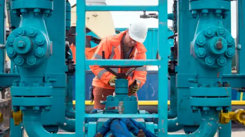 PA Media File photo from 2018 of a worker at the Cuadrilla fracking site in Preston New Road, Little Plumpton, Lancashire. The man, dressed in high-vis, orange clothes and a hard hat, is looking at part of the equipment. He is surrounded by huge blue pipes.