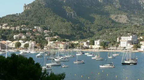 A wide shot of a bay in Majorca. There are boats and houses with a backdrop featuring trees and a large hill.