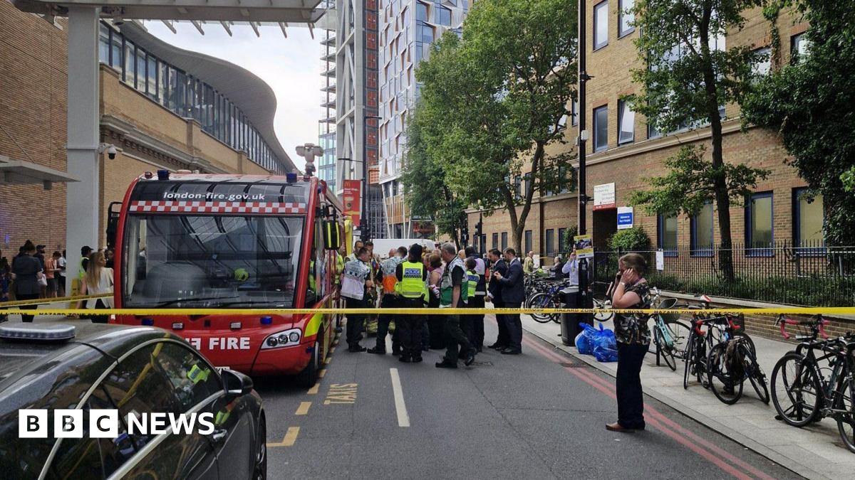 Emergency services are gathered by a fire vehicle outside a modern hospital building. There is tape closing off the road.