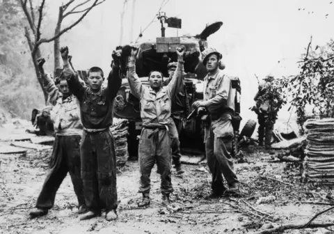 Universal History Archive/Universal Images Group via Getty Images Three Korean men in military clothing hold their hands up, with what appears to be a US solider with a gun standing behind them. His helmet is sitting at an angle on his head. Behind them is a tank, and the outline of more soldiers can be made out. The picture is black and white. 