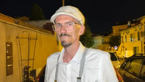 @jeanpormanove Raphaël Graven stands outside at night in the street. He is wearing a white shirt and grey suspenders, with a white flat cap. He has glasses and sharp facial hair trimmed in a type of goatee. There are cars and houses behind him lit yellow in the streetlights under the dark sky.