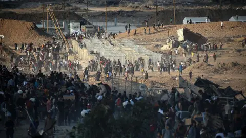 Getty Images Hundreds of people queue near metal fences on a barren, arid landscape for food. 
