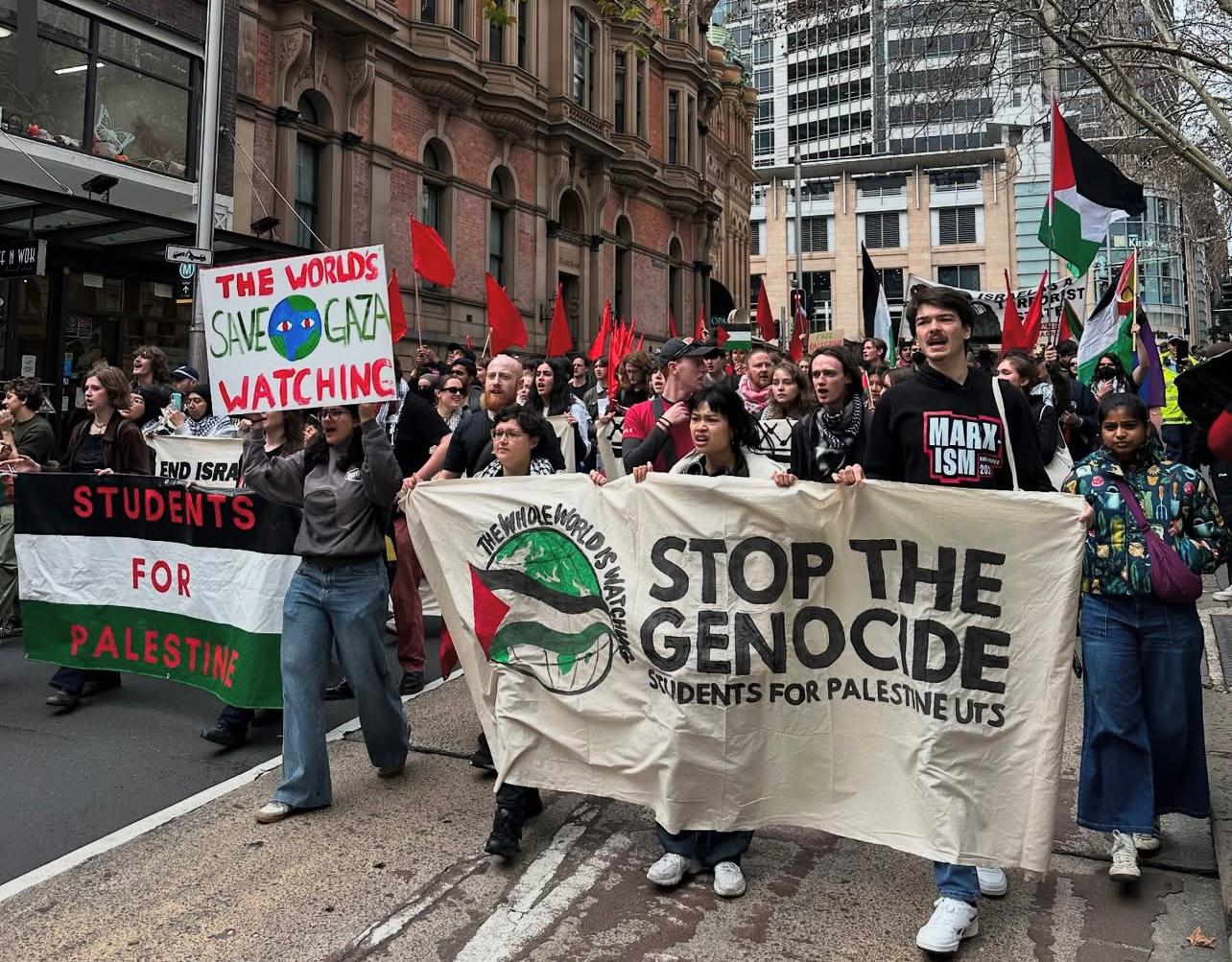 A group of protesters are marching on a city street. In the foreground, people are holding two large banners. One banner says "STUDENTS FOR PALESTINE" and the other says "THE WHOLE WORLD IS WATCHING, STOP THE GENOCIDE". A person in the middle holds a smaller sign that reads "THE WORLD'S WATCHING, SAVE GAZA". Many other people are visible behind the banners, with some holding Palestinian flags and other signs. The buildings on either side of the street suggest an urban environment.