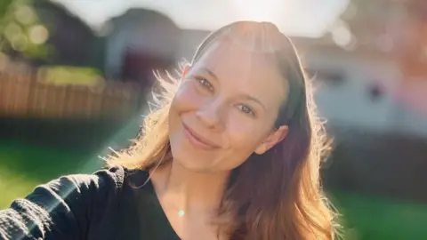 @bindisueirwin / Instagram Bindi Irwin, smiling at camera, with shoulder-length brown hair, wearing a black top and sun glare above 