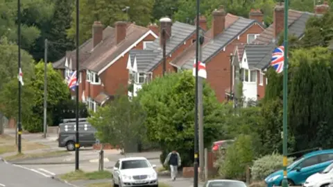 Rows of houses with England flags flying on lampposts outside. A number of parked cars can be seen outside the homes and on the pavement. A pedestrian can also be seen walking on the pavement.
