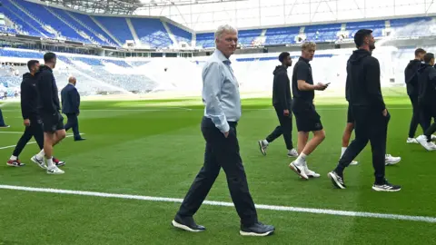 Reuters Side view as David Moyes, smiling in the centre and turning to camera, and about eight Everton players in front and behind him - who are in dark training gear - stroll around the green pitch before the game. Empty blue seats can be seen in the stands behind.