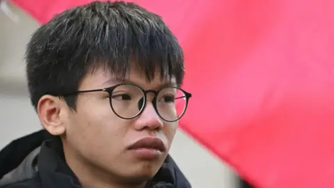 Getty Images Tony Chung, in black-rimmed glasses and a black winter jacket, attends a demonstration in London to protest the introduction of the Article 23 National Security Law in Hong Kong. Part of the Hong Kong flag is seen behind him