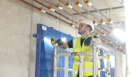 Will Cunnigham, a man in a white hard hat and hi-viz tabard, looks at a wall while working. He has a brown thin beard and moustache