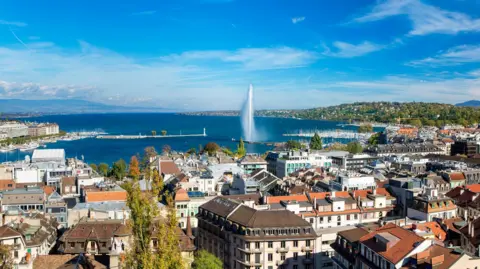 Getty Images Geneva skyline cityscape with an aerial view of Jet d'eau fountain, Lake Leman, bay and harbour from the bell tower of Saint-Pierre Cathedral. Sunny day blue sky.

