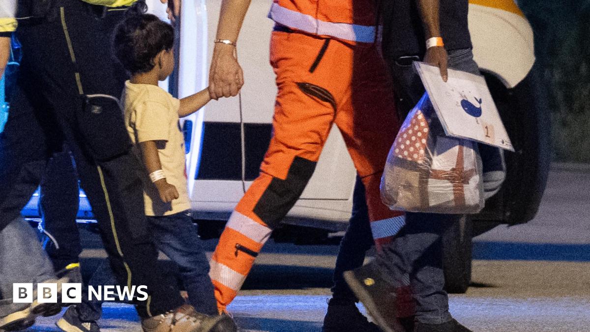 A Gazan boy being escorted off of a plane by Italian authorities. A man in an orange uniform is holding his hand and belongings
