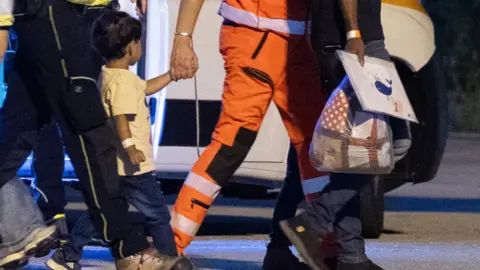 Andrea Fasani/ EPA A Gazan boy being escorted off of a plane by Italian authorities. A man in an orange uniform is holding his hand and belongings 