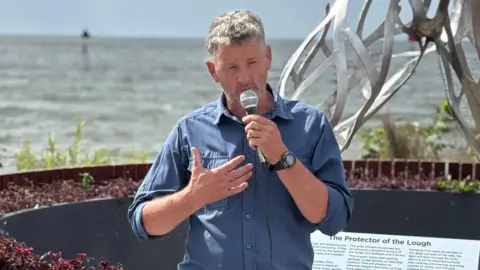 BBC A grey haired man wearing a blue shirt is speaking into a microphone on the shore of Lough Neagh