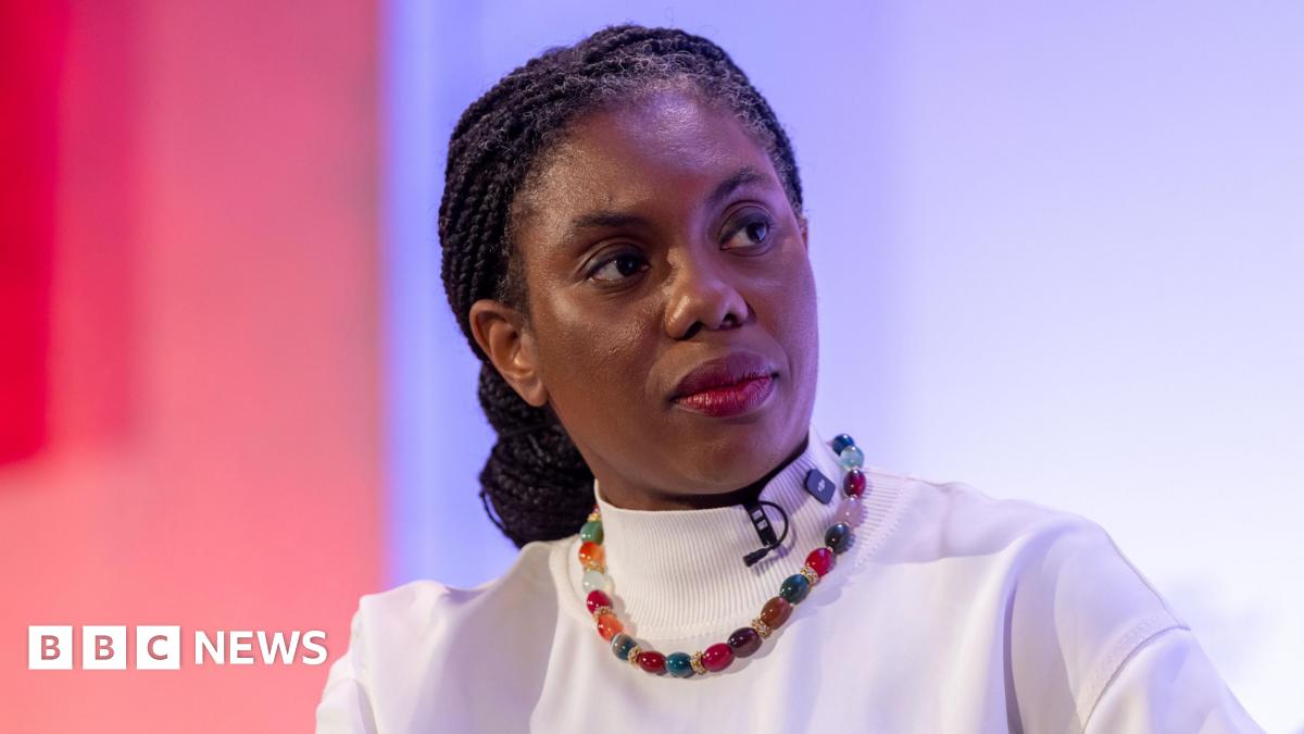 Kemi Badenoch wears a white shirt and multicoloured necklace. There is a microphone clipped to her neckline and a red and purple background behind her.