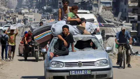 Reuters Three children on top of a battered car - one wearing black an sitting on the bonnet, the other two - one wearing a green top the other a white vest - sit on top of bed clothes on the roof. A man on a bicycle is to the left, with another on a motorcycle and yet others carrying boxes and mattresses along a stretch of road with ruins all over.
