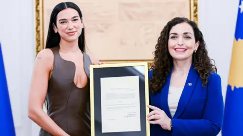 President of the Republic of Kosovo Lipa and the Kosovo president smiling holding a citizenship certificate that is in a gold frame with a black background