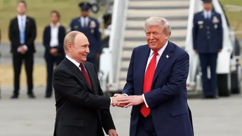 Reuters Trump and Putin stand on the tarmac in Alaska ahead of their summit, shaking hands and smiling. Behind them stand a row of uniformed men and women, and a large staircase leading up towards an aircraft. Putin is wearing a black suit, white shirt and dark red tie, and Trump is wearing a navy blue suit, white shirt and red tie. 
