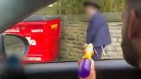 BBC Still of a man in a car spraying a water pistol at a boy walking past who wears an Orthodox Jewish hat. 