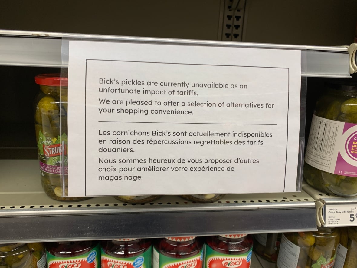A sign sits on a shelf in a grocery store in front of jars of pickles.