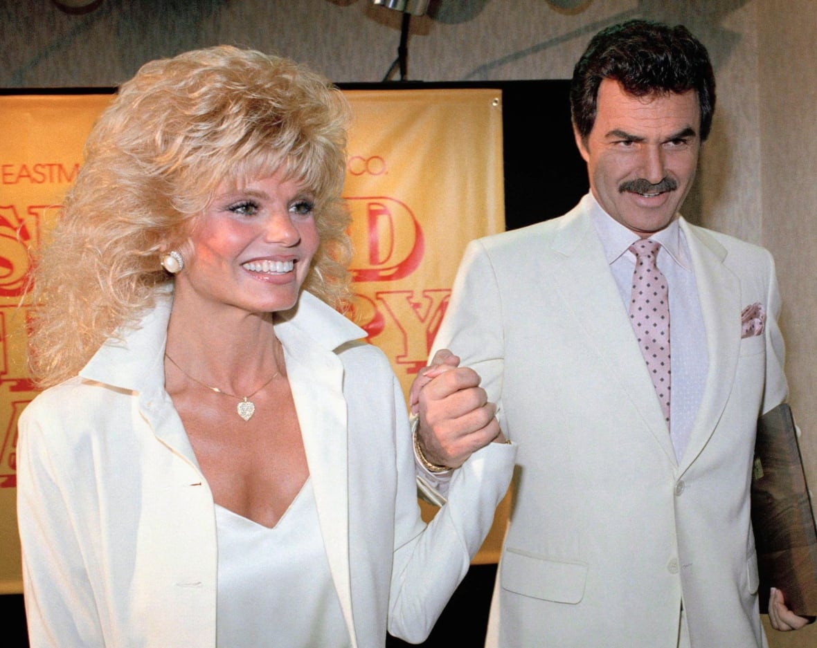 A man and woman wearing light coloured suits hold hands
