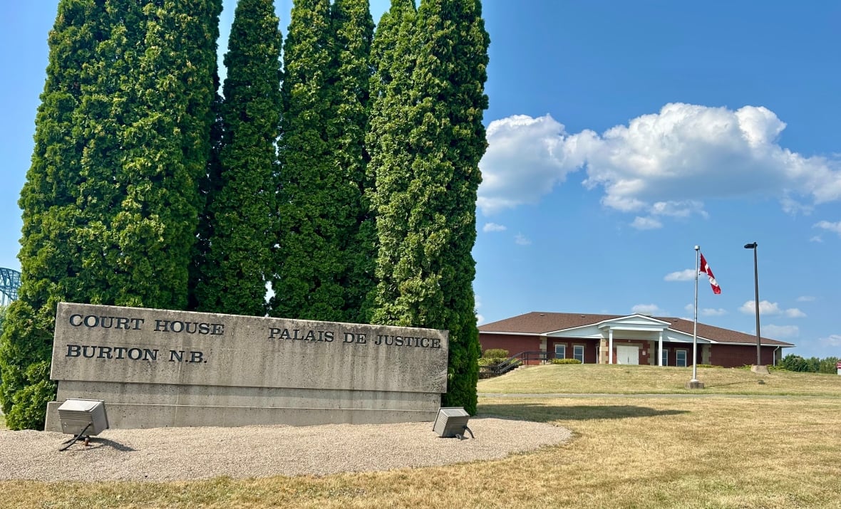 A sign reads 'Court House, Burton N.B.' in front of some tall trees with a brick building off in the background on a sunny summer day with dry grass. 