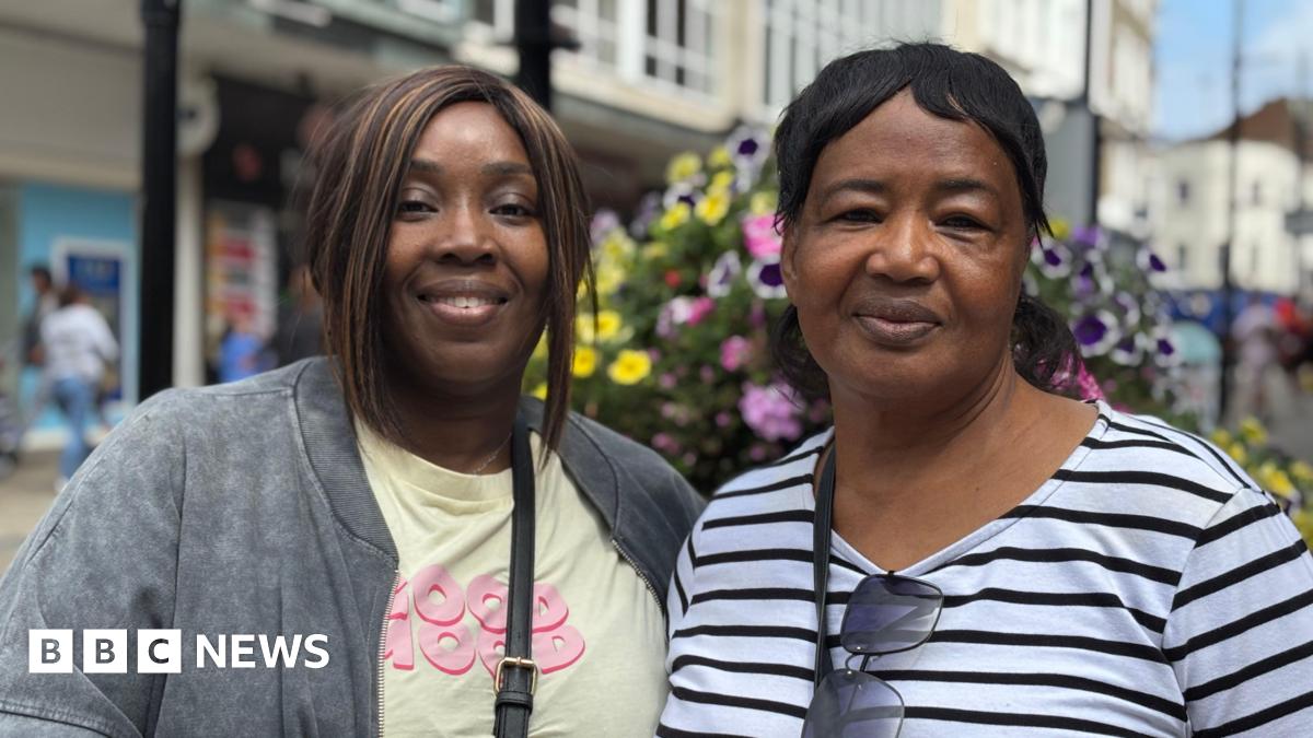 Two women - one in a blue and white striped long sleeved top with a handbag strap over her shoulder and sunglasses hanging off her collar, and the other in a yellow T-shirt and grey jacket with a crossbody bag strap visible, are standing on a high street and smiling at the camera. There are shops, floral displays and people blurred in the background.