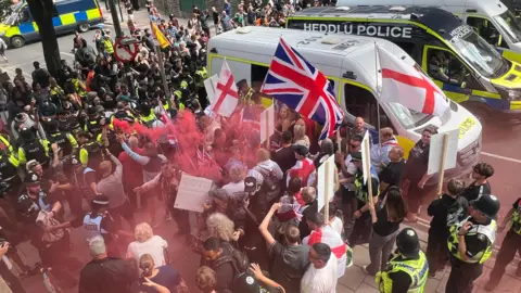 BBC Anti-immigrations protesters waving St George and union jack flags are on one side of a line of police officers on a street. One person is holding a red flare, leaving red smoke above some people's heads. On the other side are counter protesters.  There are three police vans to the side of them; the middle one says Heddlu Police and is black.
