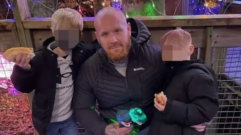 Lancashire Police Lenny Scott, who is bald and has a ginger beard, poses with his arms around his young sons. They are eating hot-dogs and their faces have been blurred.
