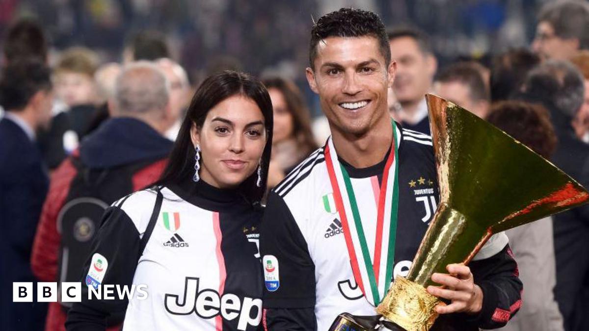 Cristiano Ronaldo is pictured wearing a medal on a red, green and white striped ribbon, and holding a large golden trophy. To his left is a woman in a matching football top, with long dark hair and silver earrings that come to her shoulders.