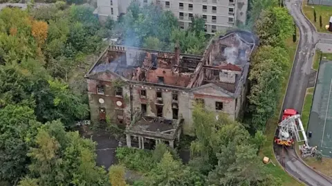 PA Media An aerial shot of Woolton Manor. It is an old house surrounded by trees. The house is still smoking and the roof has been completely destroyed.