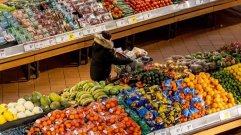 Reuters A woman in a dark coat and seen from above pushes a shopping cart through the produce section of a grocery store. On either side of her are piles of brightly coloured produce.