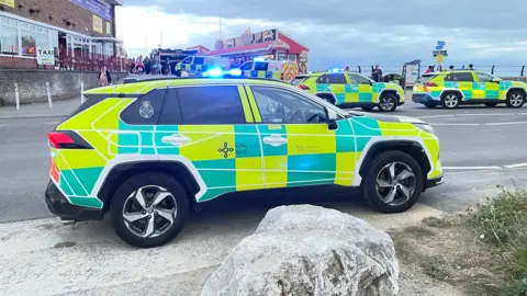An emergency vehicle is parked with its lights flashing. Two more emergency cars can be seen on the right with a further two police vans in the background. They are parked at the entrance to Coney Beach Amusement Park with the lights from one of the parks buildings in the background.
