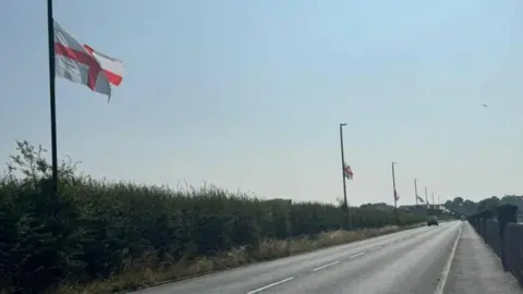 A series of lampposts have St George flags strung up on them, along the side of a main road in Birmingham. A car can be seen in the distance, and a large hedge is to the left with a wall on the right. It is a sunny day with a clear blue sky.