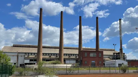 BBC/Spencer Stokes A factory with five tall narrow chimneys rising from the ground. The sky is blue with some white clouds floating above.