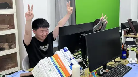 BBC Shui Zhou, a young adult who pretends to have an office job, makes the victory sign with both hands that he is holding up in the air