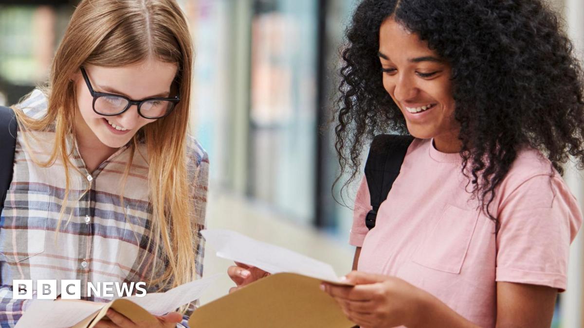 Two female students smile as they look down at exam results they've just removed from brown envelopes. The student on the left has long straight blonde hair, and wears thick-rimmed black glasses and a blue, white and red checkered shirt. The student on the right has shoulder-length dark curly hair and wears a pink t-shirt. Both students are wearing black rucksacks.