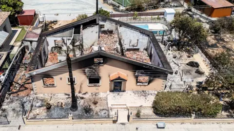 Ben Armstrong Birdseye view of the ruins which remain of the family home in Sinou, Cyprus. Burnt black grass can be seen outside of their fence on the left. The home is left without a roof, with only the house walls remaining among the rubble. 