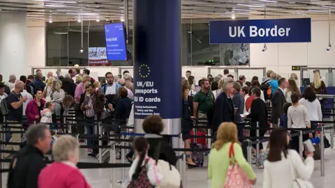 Getty Images The UK border control area at Gatwick Airport. The white-walled hall is busy with people who are queuing in a long, winding line. Some are holding suitcases and passports. There is a large blue column in the middle of the room which says: "UK/EU passports" and a large sign hangs from the ceiling which reads "UK Border".