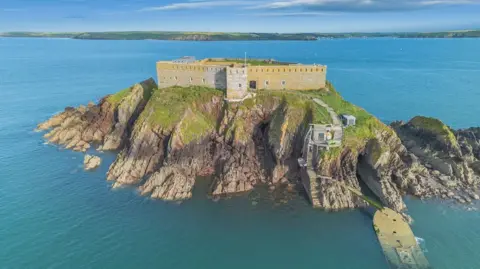Strutt & Parker An aerial view of Thorne Island, with the yellowed fortress walls encompassing it and cliffs covered in greenery at its edges. The island is surrounded by the sea, with the mainland in distance. Two flights of steps lead down to the water's edge. 
