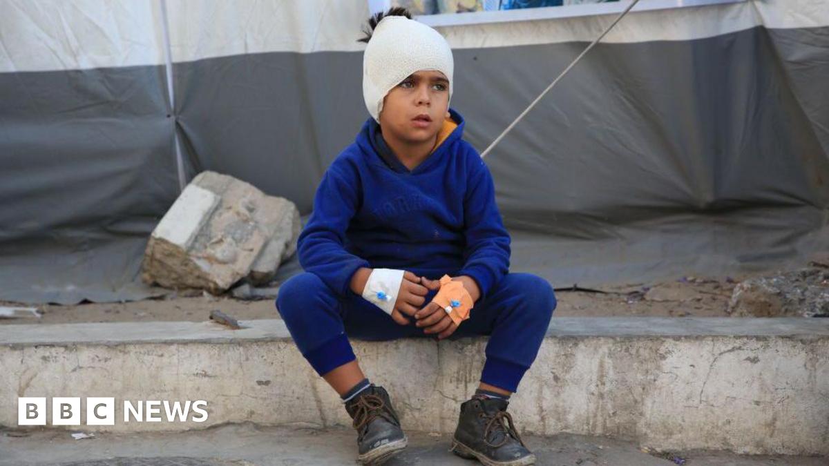 A Palestinian boy sits on the sidewalk with his head wrapped in bandages and an IV in his hands at Al-Ahli Baptist Hospital