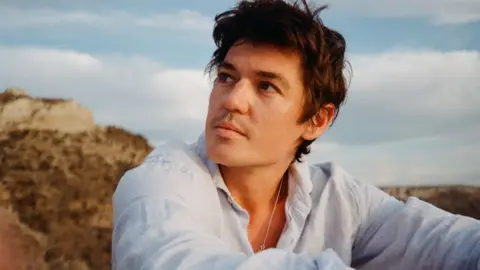 Connor James A man with brown hair in a white shirt sits outside by a grassy cliff and gazes into the distance.