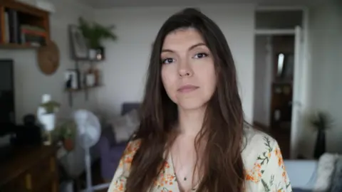 A woman in her mid 20s, with long dark hair and wearing a light yellow patterned blouse, standing in her living room gazing solemnly into the camera lens.