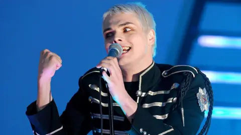 Getty Images Gerard Way performing at the 2006 MTV Video Music Awards. He has bright blonde bleached hair, and is wearing his signature Black Parade outfit which consists of a black jacket with silver/white stripes across. He's wearing slightly orange eyeshadow and is performing enthusiastically. 