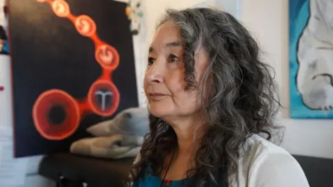 Getty Images A woman with long greying hair poses for a picture in front of a painting featuring a uterus and an IUD